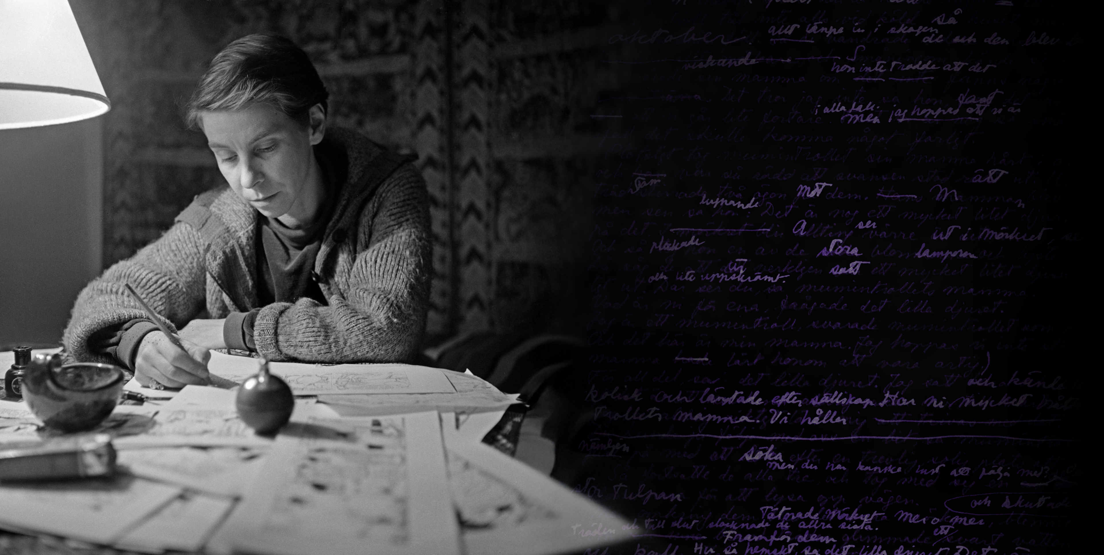 Black and white photo of a woman with short hair and a knitted sweater sitting at a desk, surrounded by papers and drawings, writing under a desk lamp.